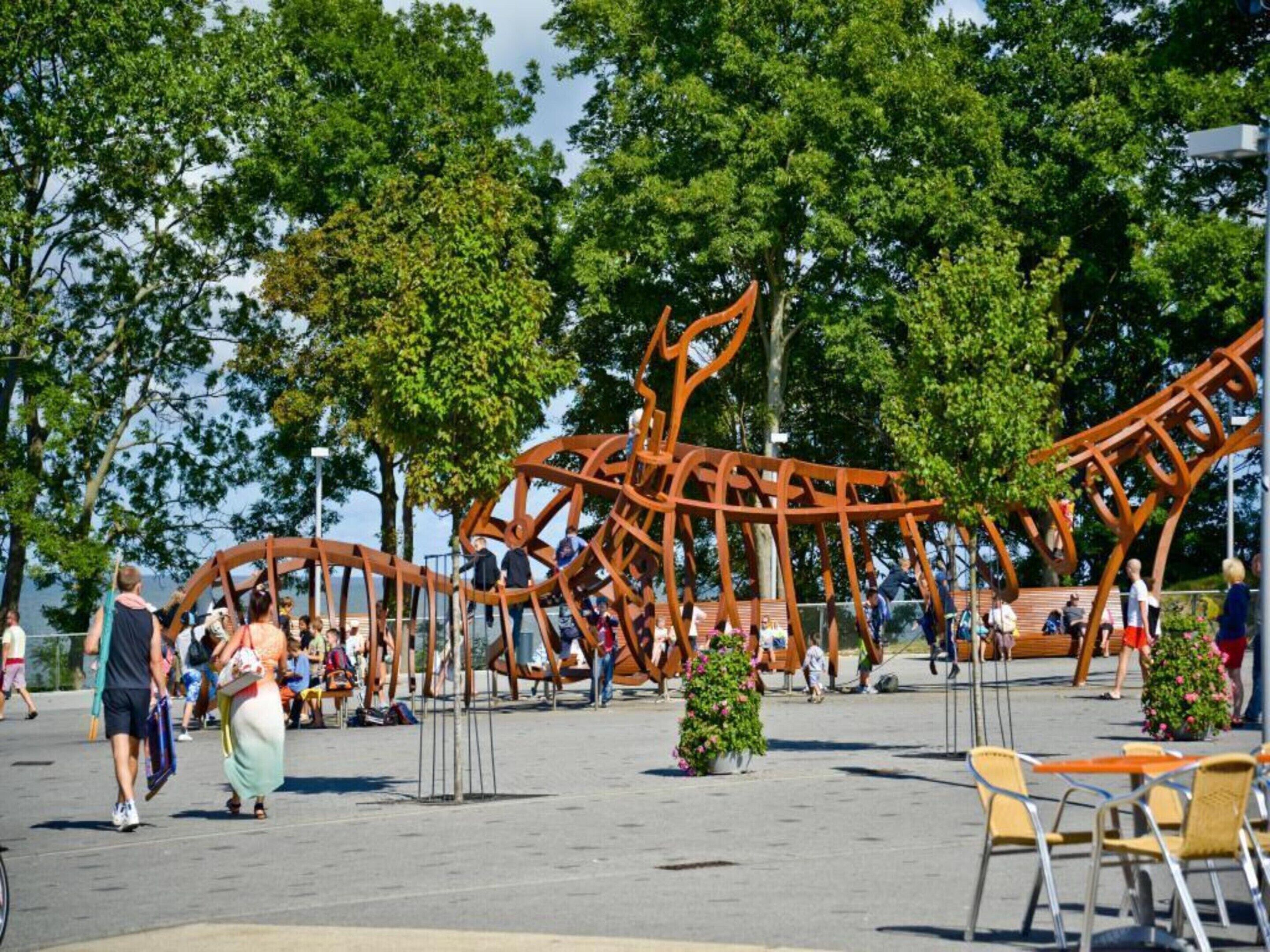 Ein öffentlicher Außenbereich mit einer großen Metallskulptur, Sitzgelegenheiten und Bäumen.