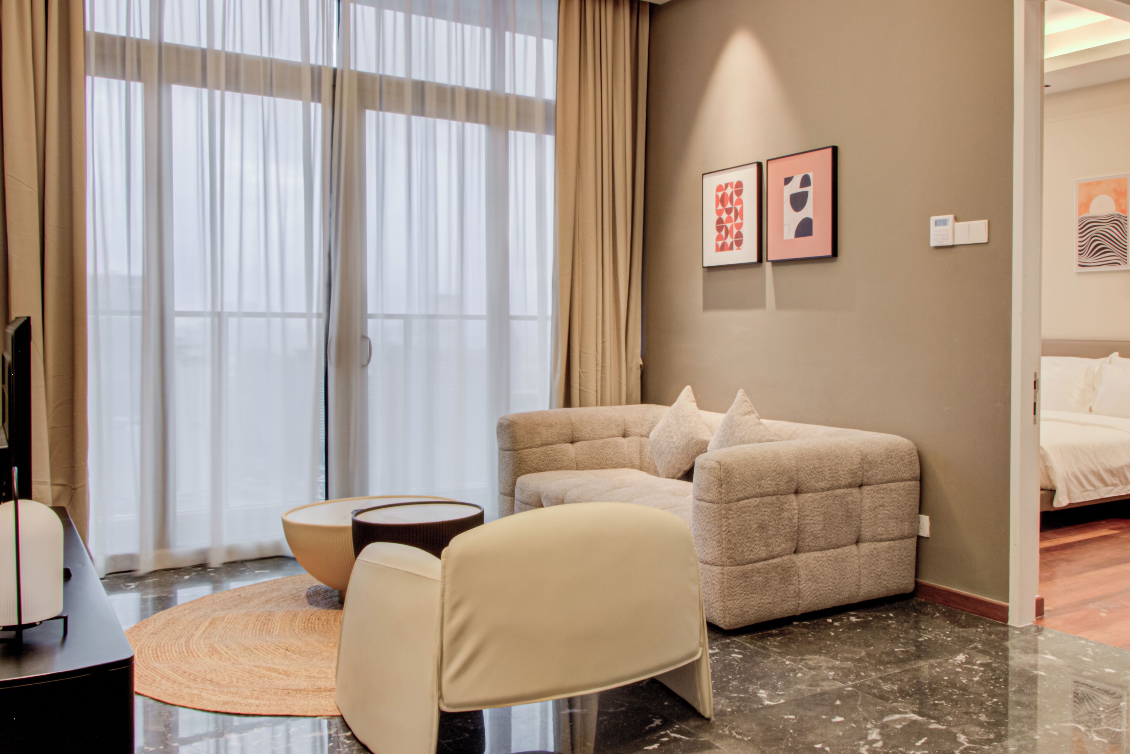 A modern hotel room with a sofa, armchair, and a round coffee table. There is a large window with curtains, a bed, and artwork on the wall.