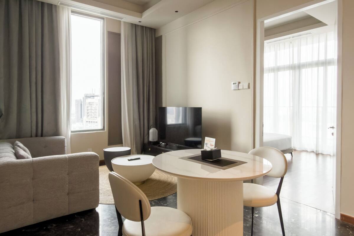 Een moderne hotelkamer met een bank, een eettafel met stoelen, een televisie en uitzicht op de stad door grote ramen.