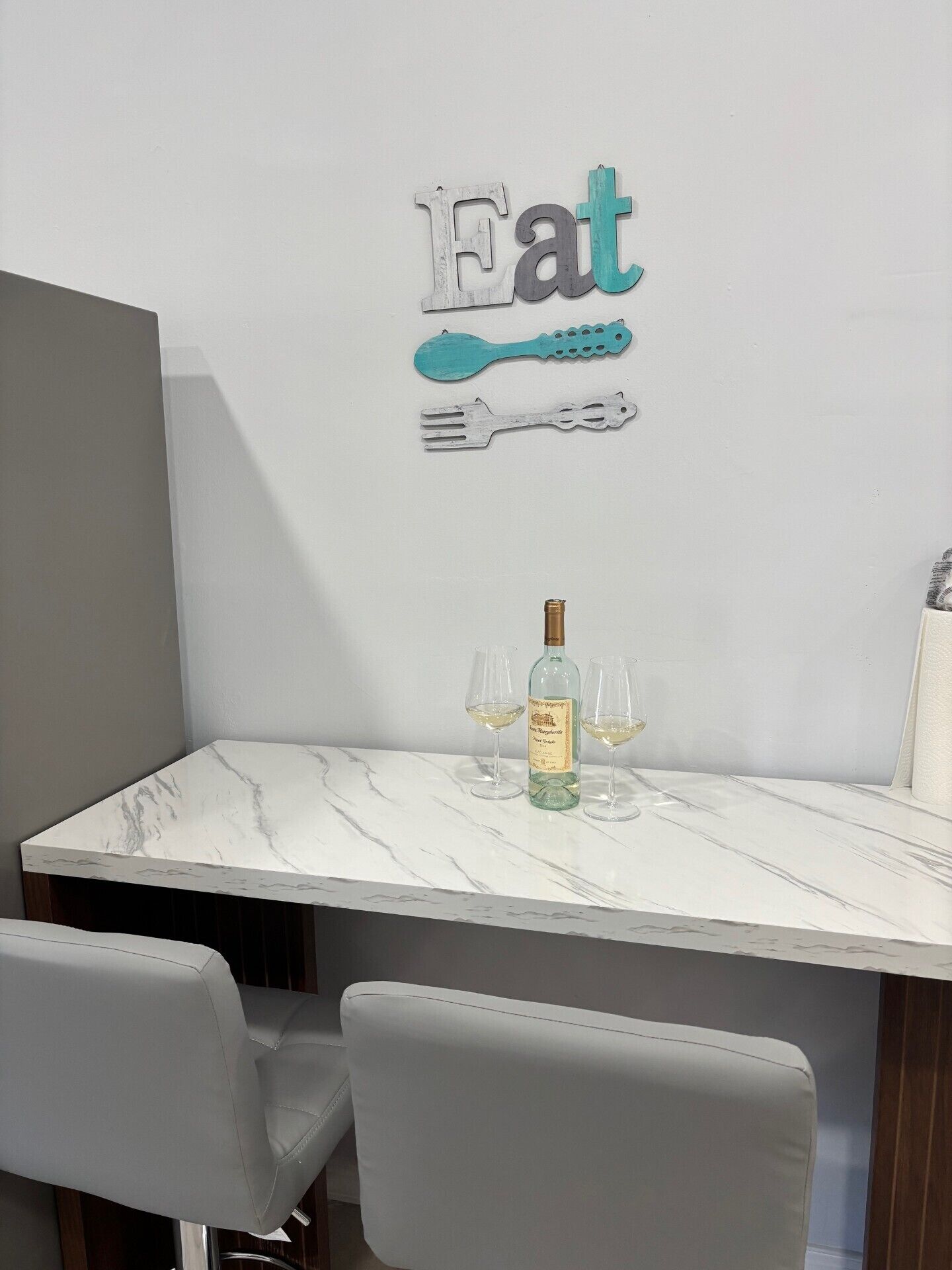 Un comedor con una mesa de mármol, dos vasos, una botella y una obra de arte en la pared que muestra la palabra 'Eat' con un tenedor y una cuchara.