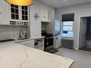 Een moderne keuken met witte kasten, een marmeren aanrechtblad, een inbouwoven en een raam met jaloezieën.