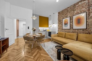 A modern living room with a herringbone floor, a sofa, a dining table with chairs, and a kitchen in the background. - Ochota Apt - Balcony, Parks Nearby - by Rentujemy (Warszawa)
