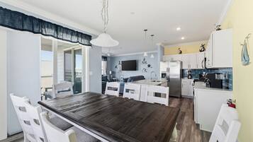 A dining area with a wooden table and white chairs, a kitchen with white cabinets, a TV mounted on the wall, and a balcony with a view.