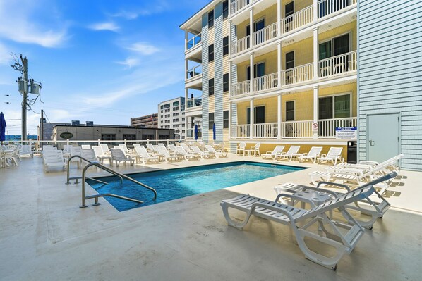 A pool area with lounge chairs, a pool, and a multi-story building with balconies.