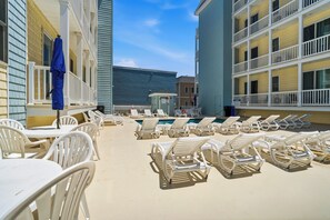 A poolside area with lounge chairs and tables, a covered seating area with a blue umbrella, and a building with multiple balconies.