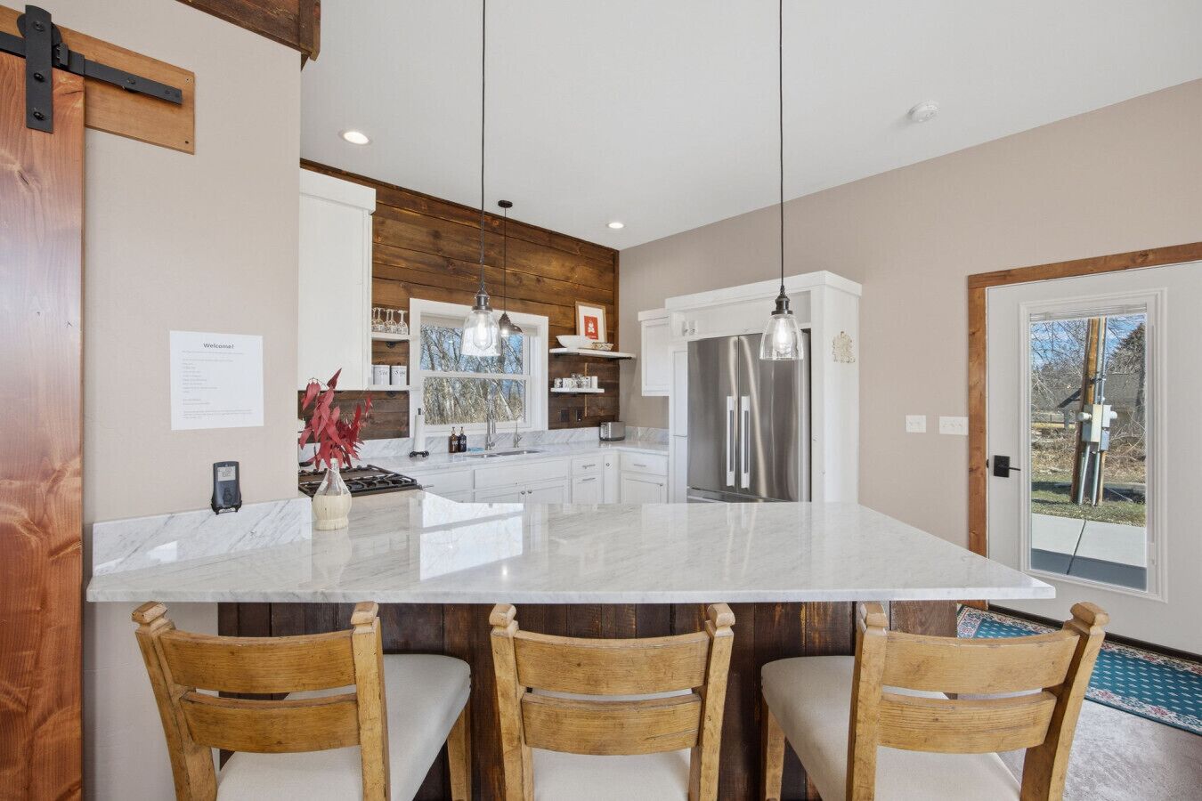 A modern kitchen with a large island, wooden chairs, stainless steel appliances, and pendant lighting.