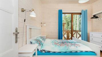 Quarto com teto de madeira, cama com roupa de cama floral, mesa de cabeceira, cómoda branca e janela com cortinas azuis.