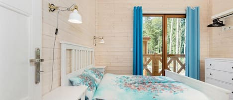 A bedroom with a wooden ceiling, a bed with floral bedding, a bedside table, a nightstand, a white dresser, and a window with blue curtains.