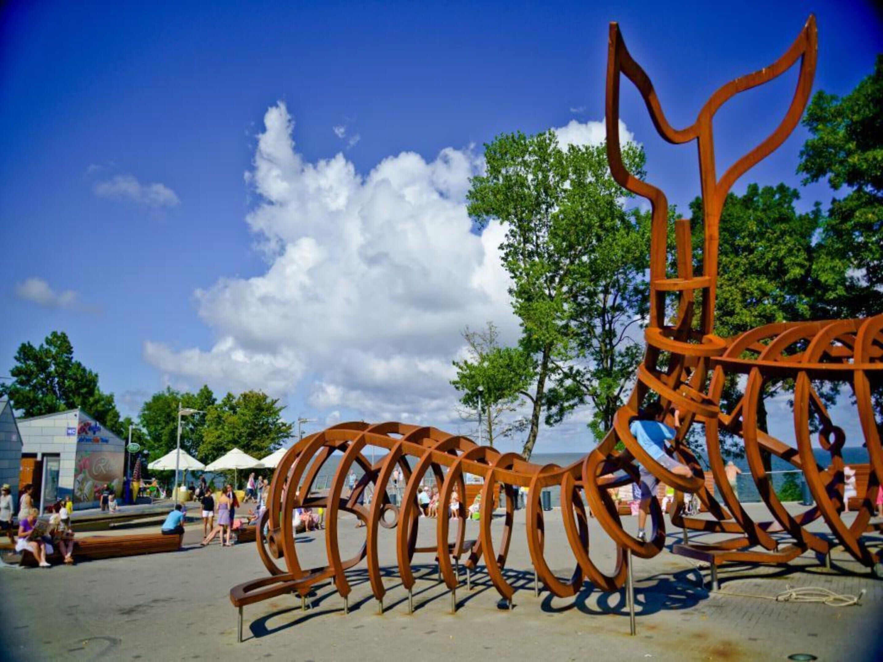 Eine Metallskulptur in einem öffentlichen Außenbereich, im Hintergrund Bäume und ein klarer Himmel.