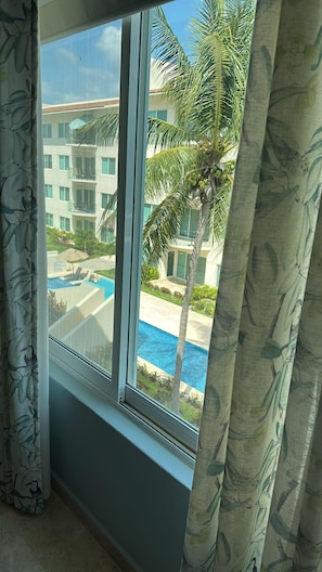 A window with a view of a swimming pool, palm trees, and a building with multiple windows. - Private and very comfortable apartment, ideal for couples or families. (Ixtapa Zihuatanejo)