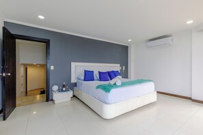 Ein modernes Schlafzimmer mit einem großen Bett, einem weißen Kopfteil und blauen Kissen. Ein Nachttisch mit Telefon und einer Wasserflasche steht bereit. Die Wand ist grau und die Decke weiß mit eingelassener Beleuchtung.