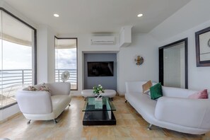 A modern living room with a flat-screen TV, white sofas, a glass coffee table, and a balcony with a view. - Oceanfront Triplex Penthouse: 4 Bedrooms (Cartagena de Indias)