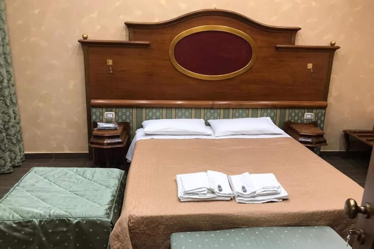 Habitación de hotel con una cama grande, dos mesitas de noche, un banco tapizado en verde y un cabecero de madera con espejo.