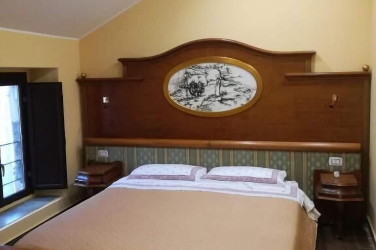 Una habitación de hotel con una cama, cabecera de madera con un plato decorativo, una mesita de noche y una ventana.