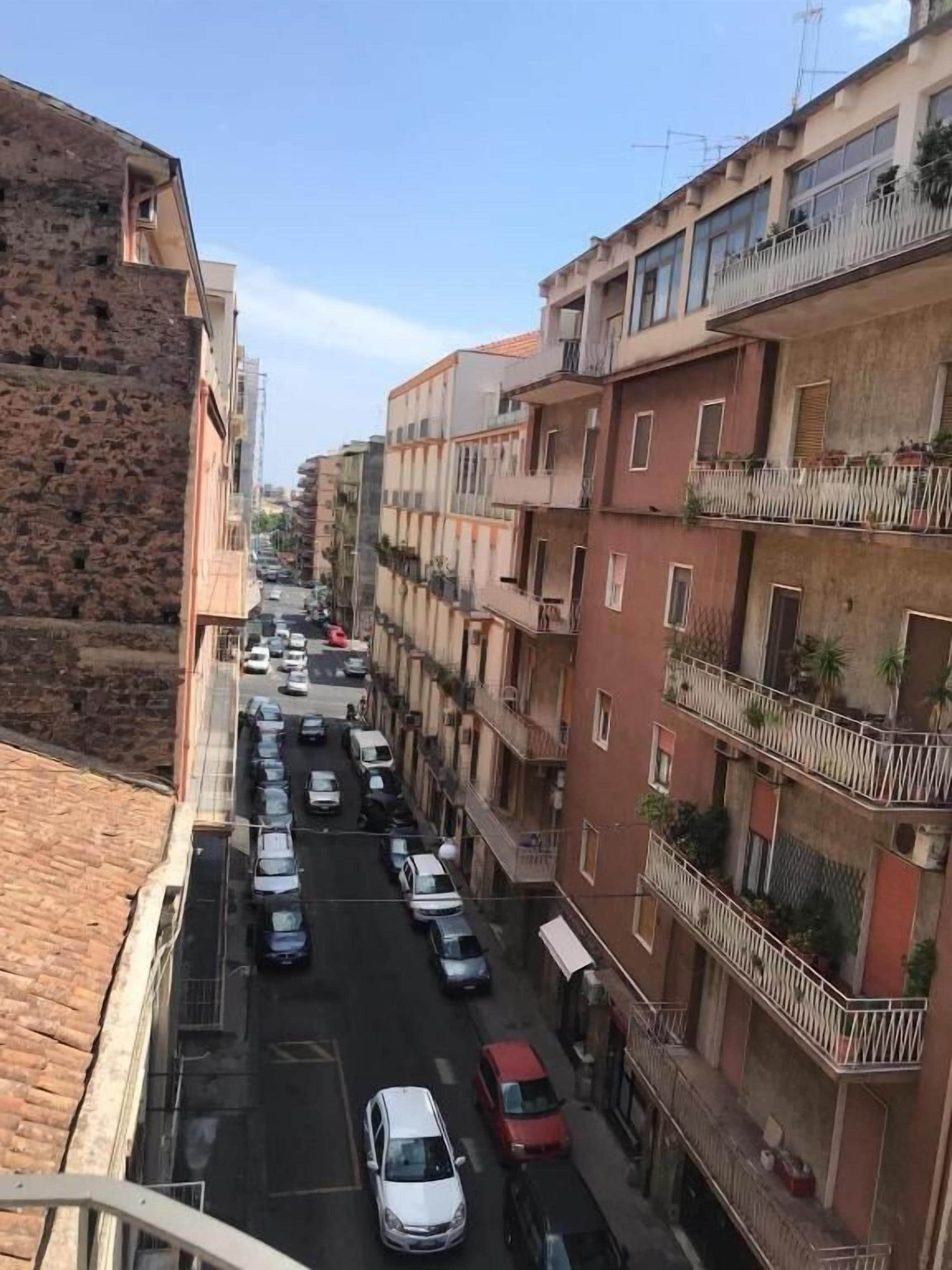 A narrow street lined with multi-story residential buildings, each with balconies and a row of parked cars.