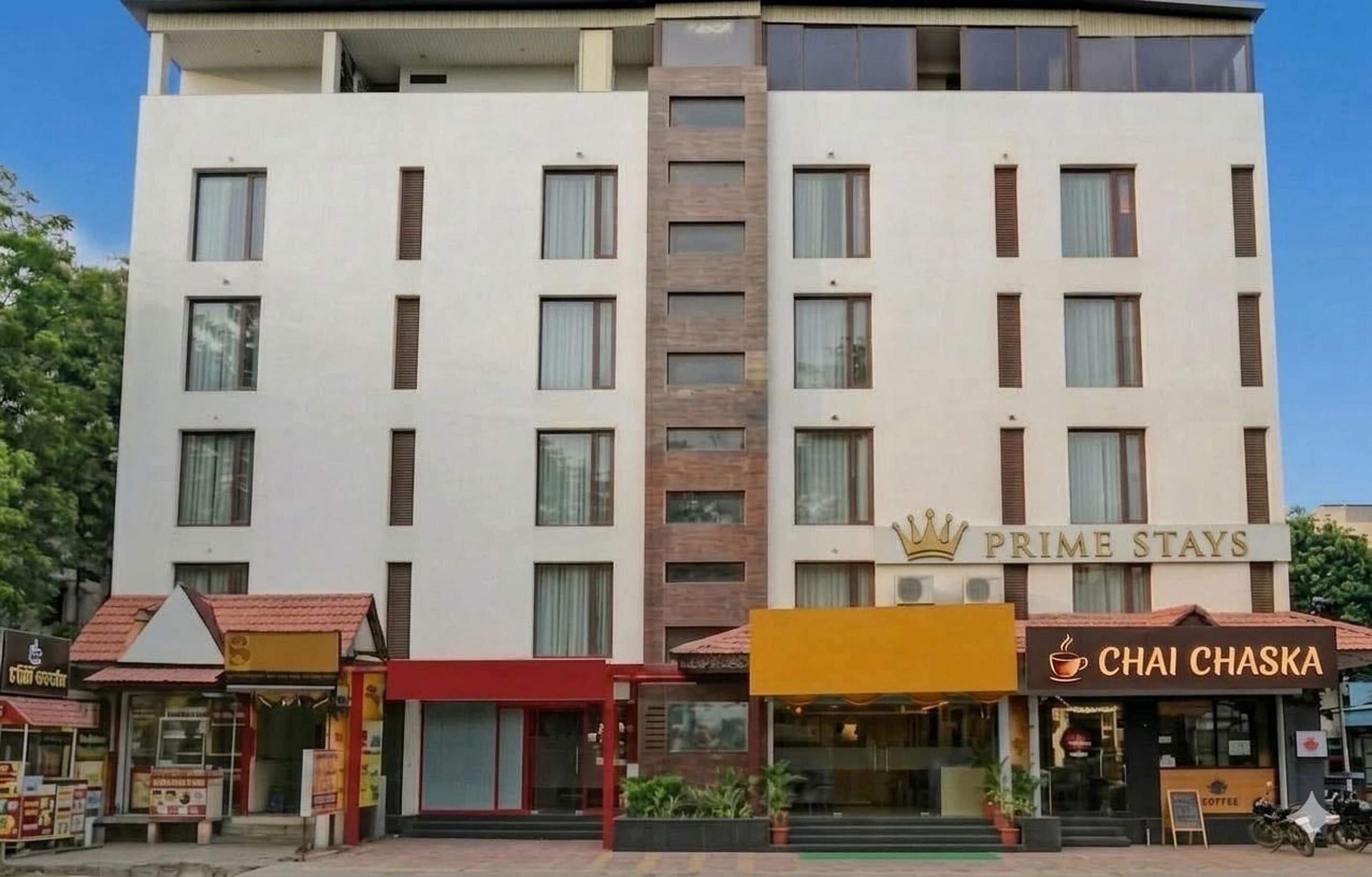 A hotel named 'Prime Stays' with a modern design, featuring a central brown column, multiple windows, and a sign for 'Chai Chaska' café.