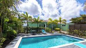 Une piscine avec une terrasse carrelée, entourée de palmiers et une maison avec un toit en tuiles.