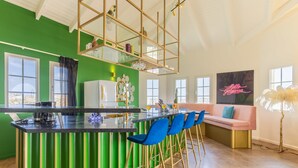 Une cuisine moderne avec un mur d’accent vert, un coin bar avec des chaises bleues, un canapé rose et un panneau indiquant « Bonjour, beauté ! ».