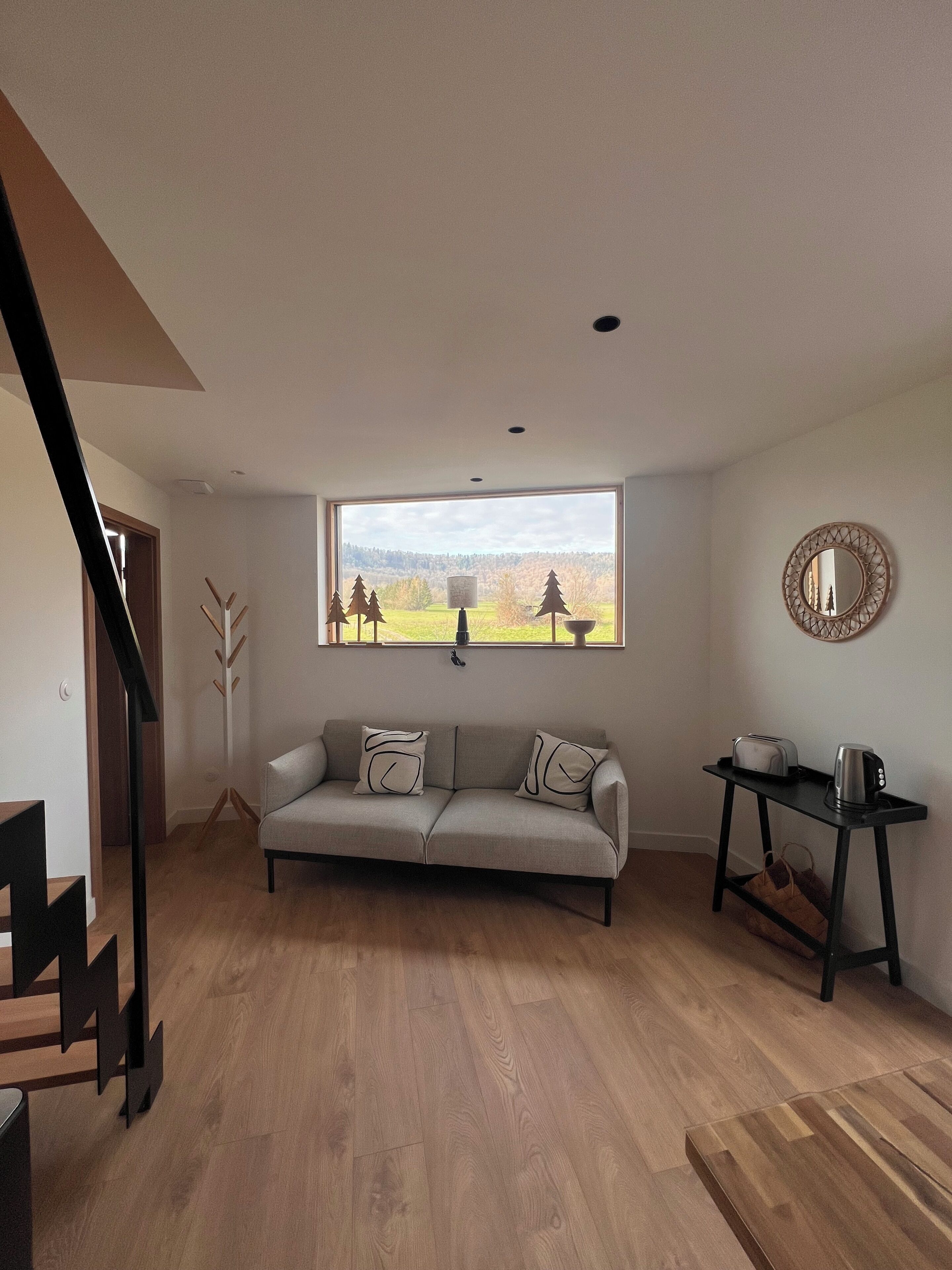 A modern living room with a sofa, a coffee table, a view of a forest, and a staircase.