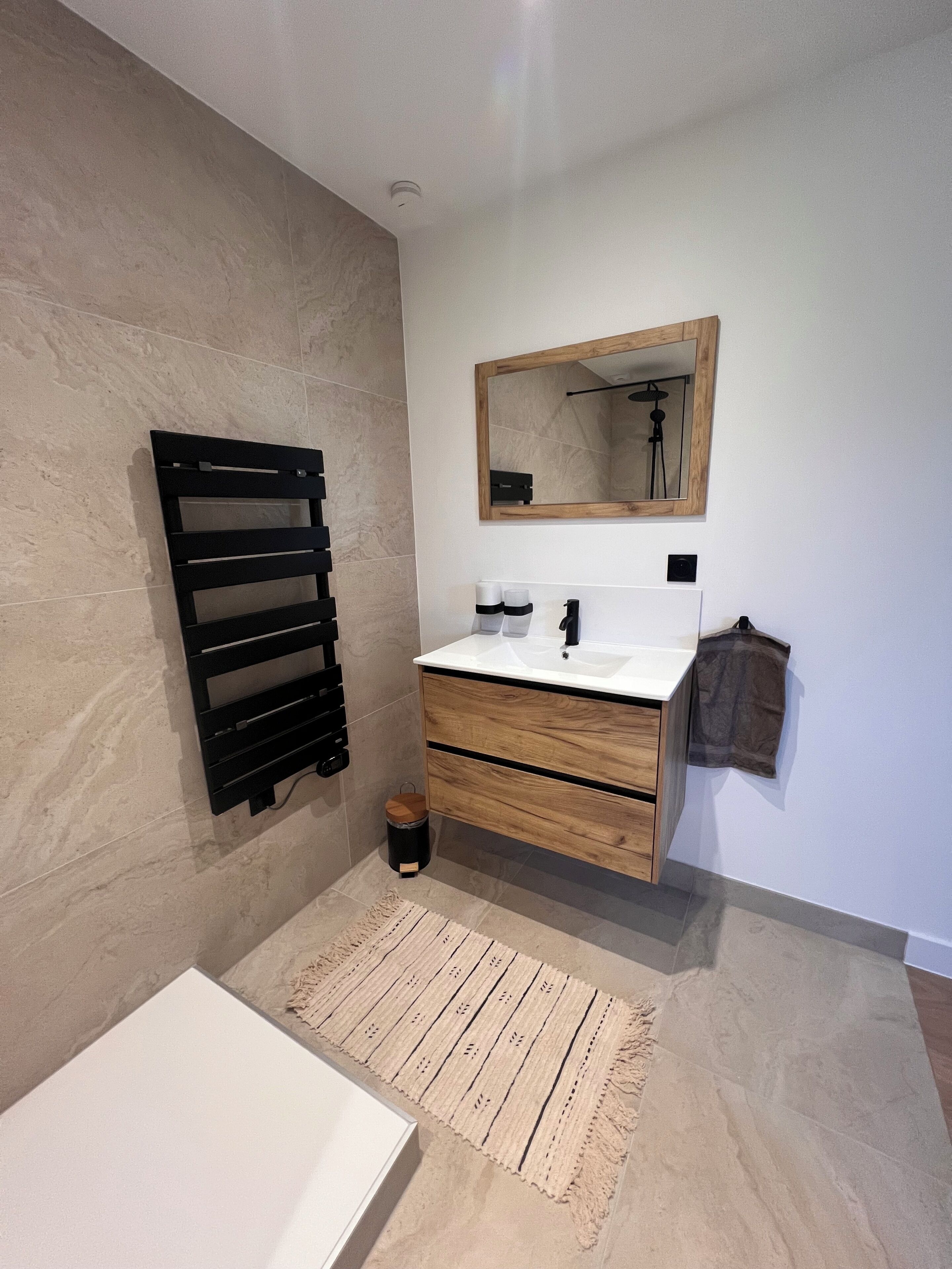 Une salle de bain avec un lavabo blanc, un meuble-lavabo en bois, un miroir, un porte-serviettes et une petite poubelle.