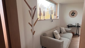 Ett modernt vardagsrum med en soffa, en lampa, ett soffbord och ett fönster med utsikt över träd.