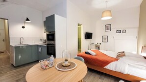 Ein kleines Hotelzimmer mit Küchenzeile, einem runden Esstisch, einem Bett mit orangefarbener Decke und einem an der Wand befestigten Fernseher.