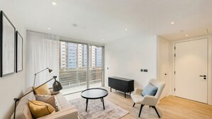 A modern living room with a sofa, a round table, a chair, a cabinet, and a large window with a view of buildings. - Refined Luxury 2B2B Home – right next to Battersea Power Station (London)