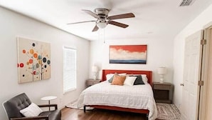 Un dormitorio con cama, una silla, ventilador de techo y obras de arte en las paredes.