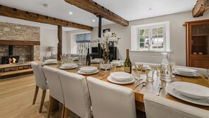 A dining area with a long wooden table set for six, surrounded by chairs, with a stone fireplace and a window in the background. - Bibury Mill - Pet friendly, Bibury (Bibury)