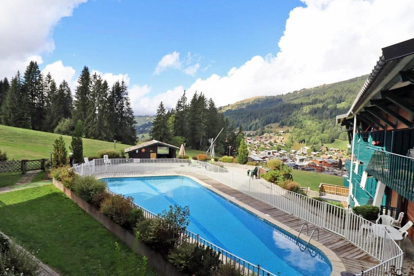 Une piscine dotée d’une terrasse en bois, entourée d’une clôture blanche, avec en toile de fond un paysage montagneux et un petit village.