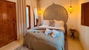 Quarto de hotel com uma cama grande, cabeceira estampada, duas luminárias fixadas na parede, uma mesinha de cabeceira pequena e um guarda-roupa de madeira.