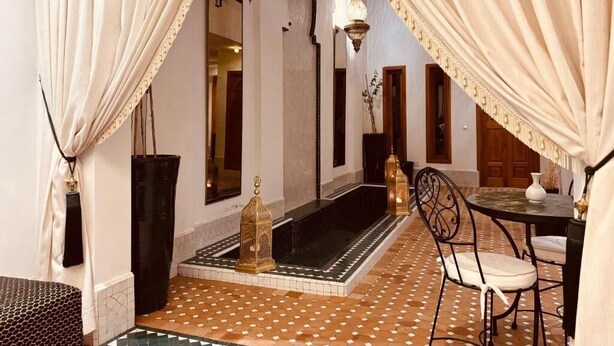 Um interior marroquino com piso de azulejos, mesa com padrão preto e branco, uma cadeira branca e um abajur preto.