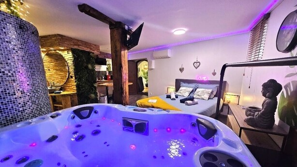 A room with a large hot tub, a bed with a headboard, a wall-mounted clock, and a small statue on a table. - Jacuzzi relaxation near Colmar (Bergheim)