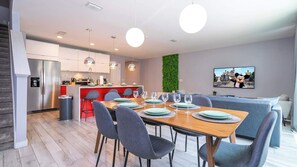 Uma cozinha moderna com frigorífico embutido, um espaço de refeições com mesa de madeira e cadeiras cinzentas, e uma sala de estar com sofá cinzento.