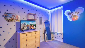 Quarto com televisão exibindo personagens animados, cômoda, espelho e adesivo de parede do Mickey Mouse.