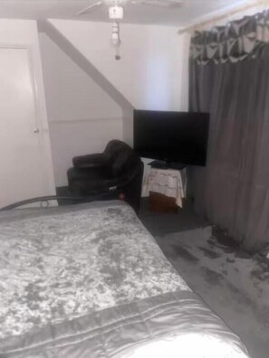 Un dormitorio con cama, televisión, una mesita pequeña y una cortina.