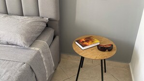 A modern room with a grey sofa, a small round wooden table with a book and glasses, and a wall-mounted speaker. - Woven Calm • 2BR Near Beach, Gateway to Chefchaouen (Martil)