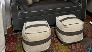 Un canapé gris foncé avec des coussins à rayures, deux poufs assortis, un tapis à motifs et une fenêtre ornée de rideaux légers.
