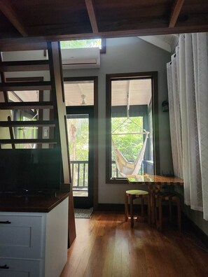 A room with a wooden floor, a TV, a hammock, and a window with a view of greenery.