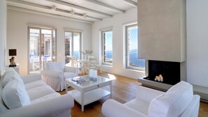 A spacious living room with white furniture, large windows, and a fireplace. - Stone Villa with Panoramic Sea Views over Mylopotas, Ios (P842+3W Chora)