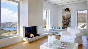 A modern living room with a fireplace, white furniture, and a view of the sea. - Stone Villa with Panoramic Sea Views over Mylopotas, Ios (P842+3W Chora)