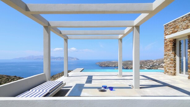 A terrace with a white pergola, a stone building, and a view of the sea and mountains. - Stone Villa with Panoramic Sea Views over Mylopotas, Ios (P842+3W Chora)