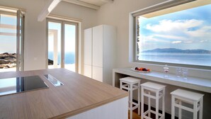 A modern kitchen with a large island, white bar stools, and a view of the sea. - Stone Villa with Panoramic Sea Views over Mylopotas, Ios (P842+3W Chora)