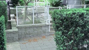 Een verhard pad dat leidt naar een hekpoort met een metalen poort, omringd door verschillende struiken en een kleine boom.