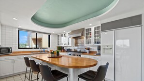 A modern kitchen with a central island, bar stools, white cabinetry, and a large window. - Lux Ocean View Penthouse 2 Kings+, Steps to Beach (Lahaina)