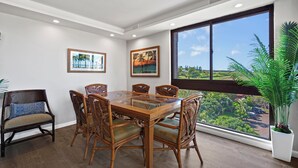 A dining area with a glass-top table, wooden chairs, and a large window with a view of greenery. - Lux Ocean View Penthouse 2 Kings+, Steps to Beach (Lahaina)