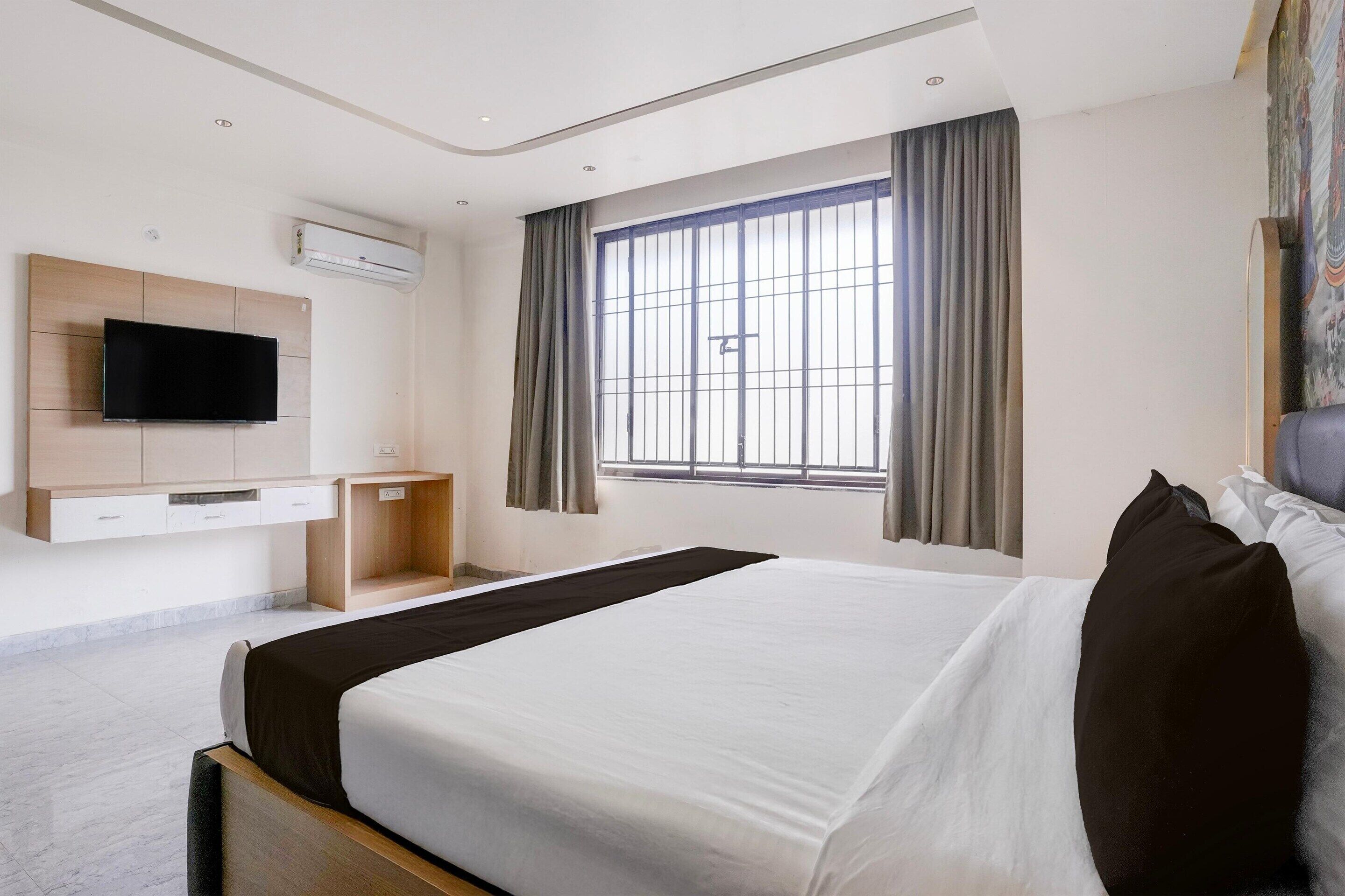 A hotel room with a bed, a TV mounted on the wall, a window with curtains, and a flat-screen TV.