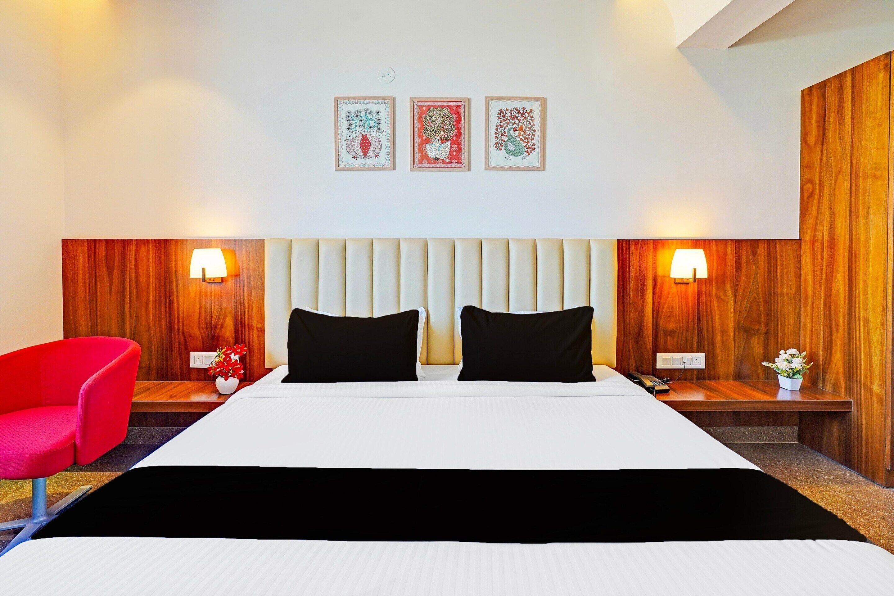 A hotel room with a bed, two bedside tables, a red chair, and three framed artworks on the wall.