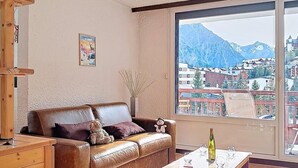 Ein Wohnzimmer mit einer Ledersofa, einem Holztisch, einer Pendelleuchte, einem großen Fenster mit Blick auf die Berge und einem kleinen Tisch mit einer Flasche und Gläsern.