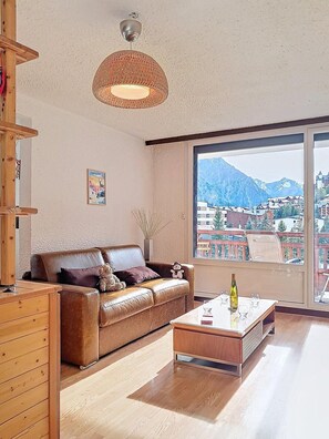 Ein Wohnzimmer mit einer Ledersofa, einem Holztisch, einer Pendelleuchte, einem großen Fenster mit Blick auf die Berge und einem kleinen Tisch mit einer Flasche und Gläsern.